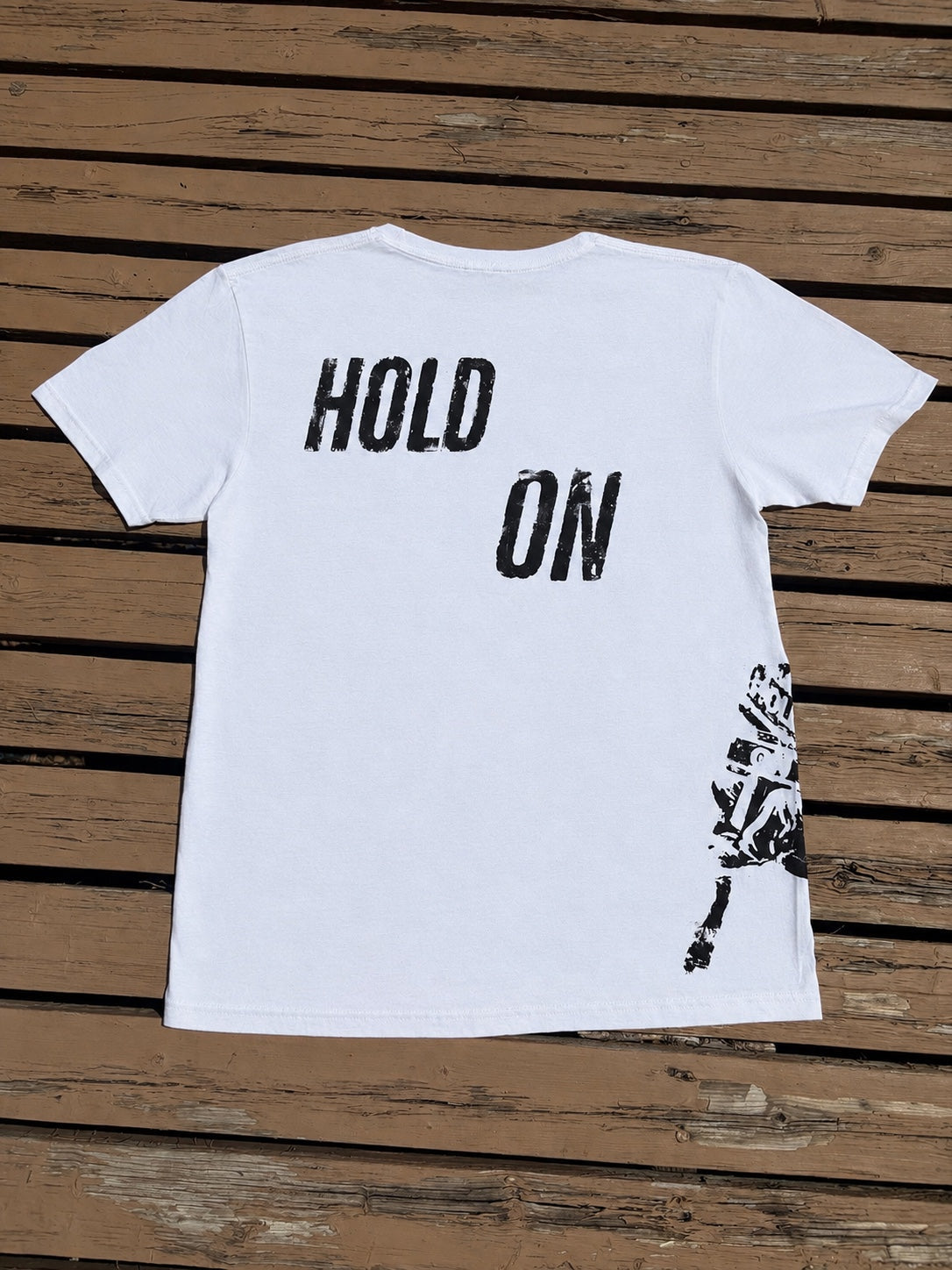 Back of white Hold On graphic t-shirt laid flat on wooden deck