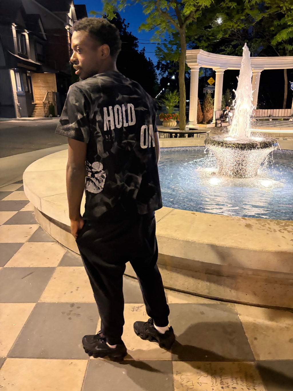 Back view of model wearing Hold On Shadow Graphic Tee by fountain at night